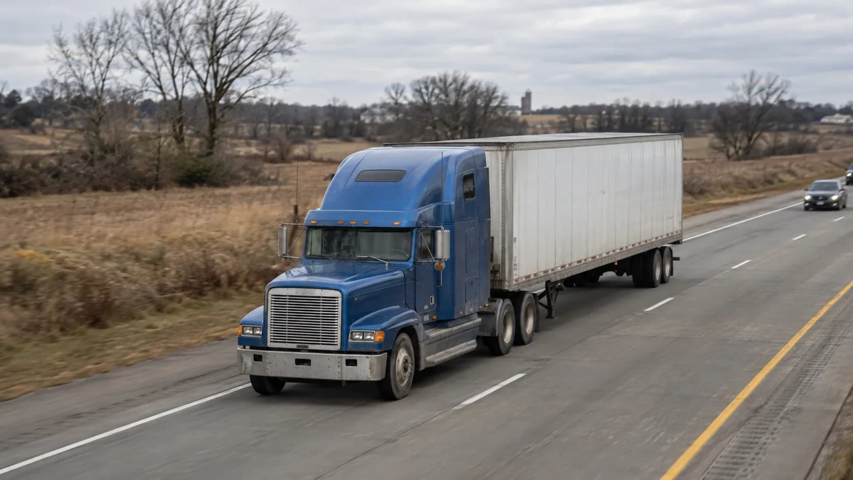 A blue aero-hood semi tractor with a mid-height sleeper pulling a white dry van trailer on a highway through winter farmland with bare trees and a silo.