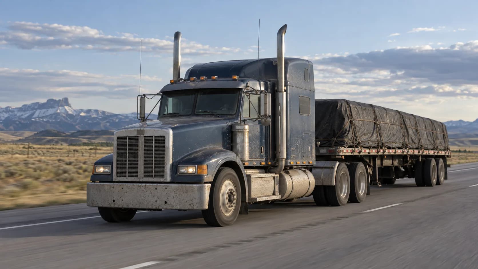Dusty blue sloped-hood rig, tarped flatbed, snowy peaks behind it. Back in the late 80s, the aero nose was the new look on the interstate. Remember when?