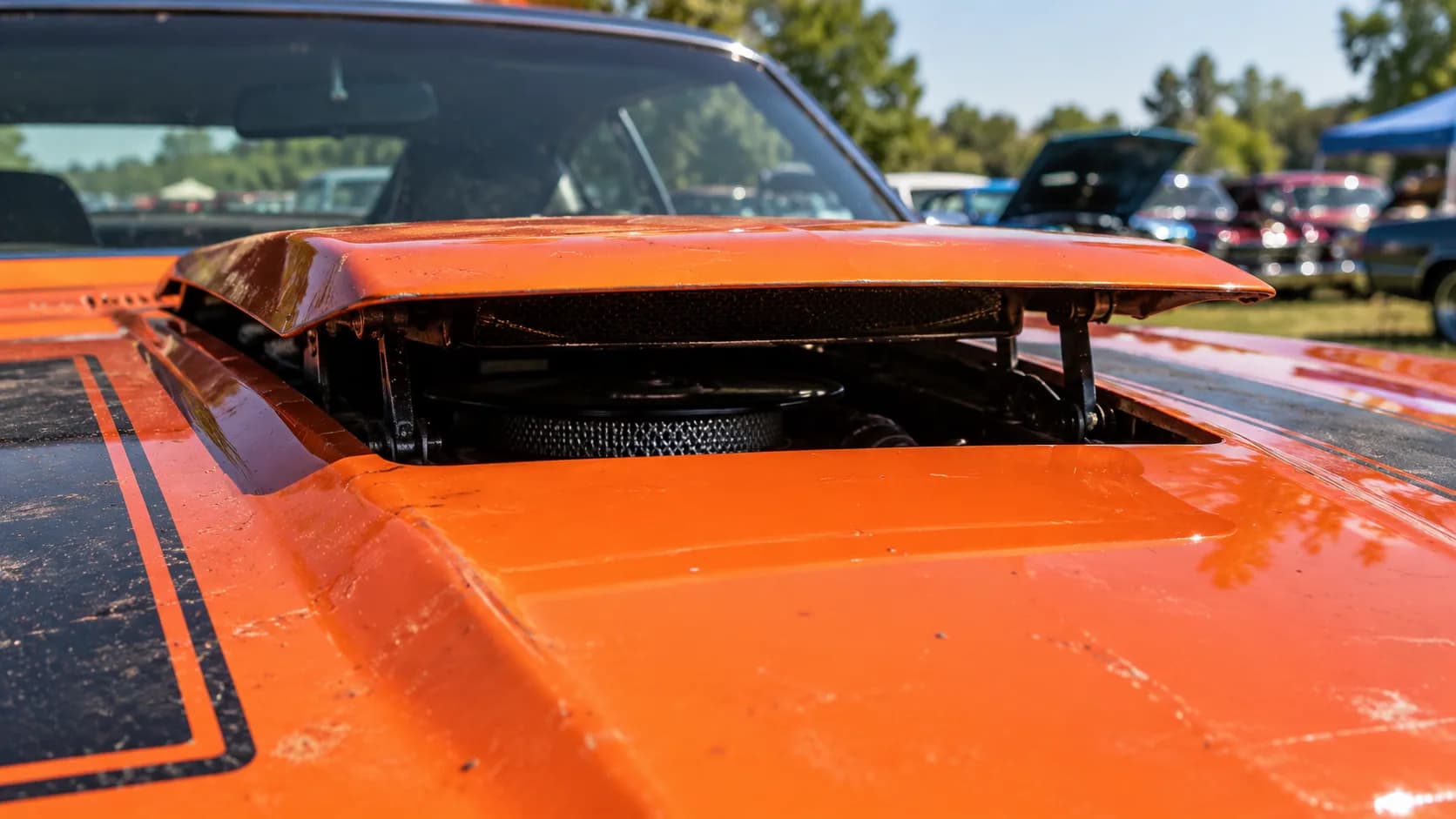 A hood scoop flipped up to show the air cleaner underneath, orange paint baking at a summer car show. In 1970, Plymouth let Road Runner drivers do that with a switch on the dash.